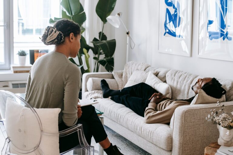 Ethnic female therapist with clipboard sitting near male client lying on couch with clasped hands during psychotherapy session in cozy office