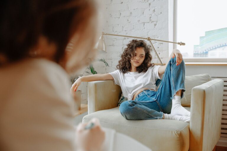 A young woman sits on a sofa during a therapy session, looking contemplative.
